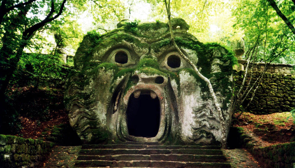 Uno dei mascheroni immerso nel bosco, fatto in pietra grigia spalanca la bocca in cima alle scale quasi come a mangiare l'ospite