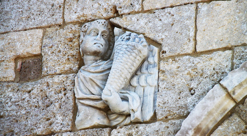 particolare di un facciata di Sant'Antimo dove emerge fra le pietre la testa scolpita di un personaggio